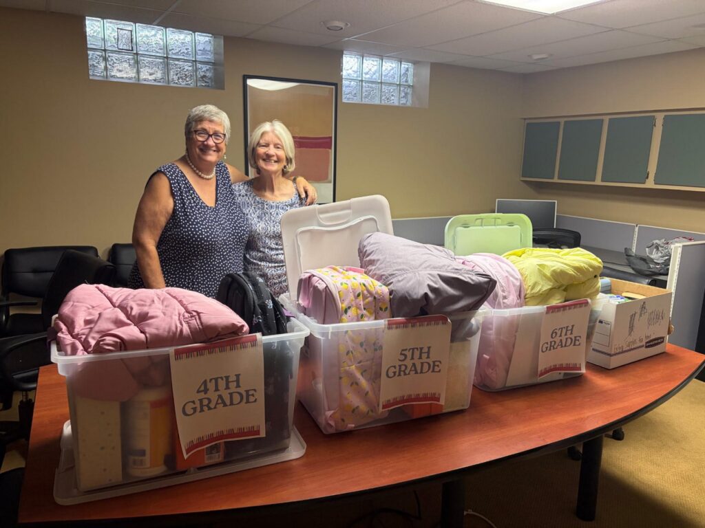 Two Grace in Action volunteers with bins of school supplies and coats sorted by grade