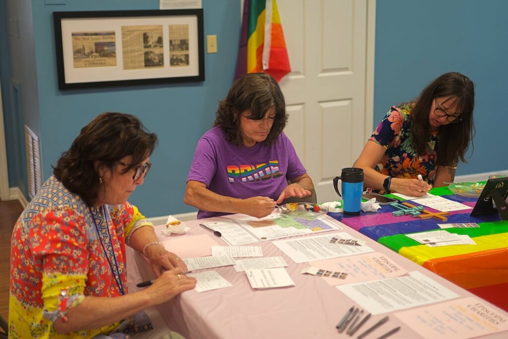 Parishioners writing postcards at a Pride celebration after Pride Mass, with a Progress Pride flag on the wall